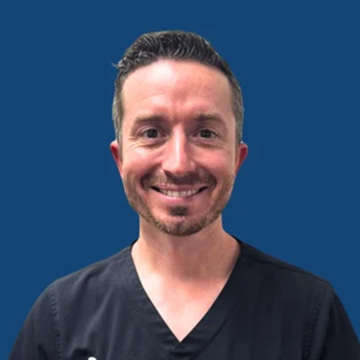 Chiropractor Zachary S. Cadman Smiling In Front Of Blue Background